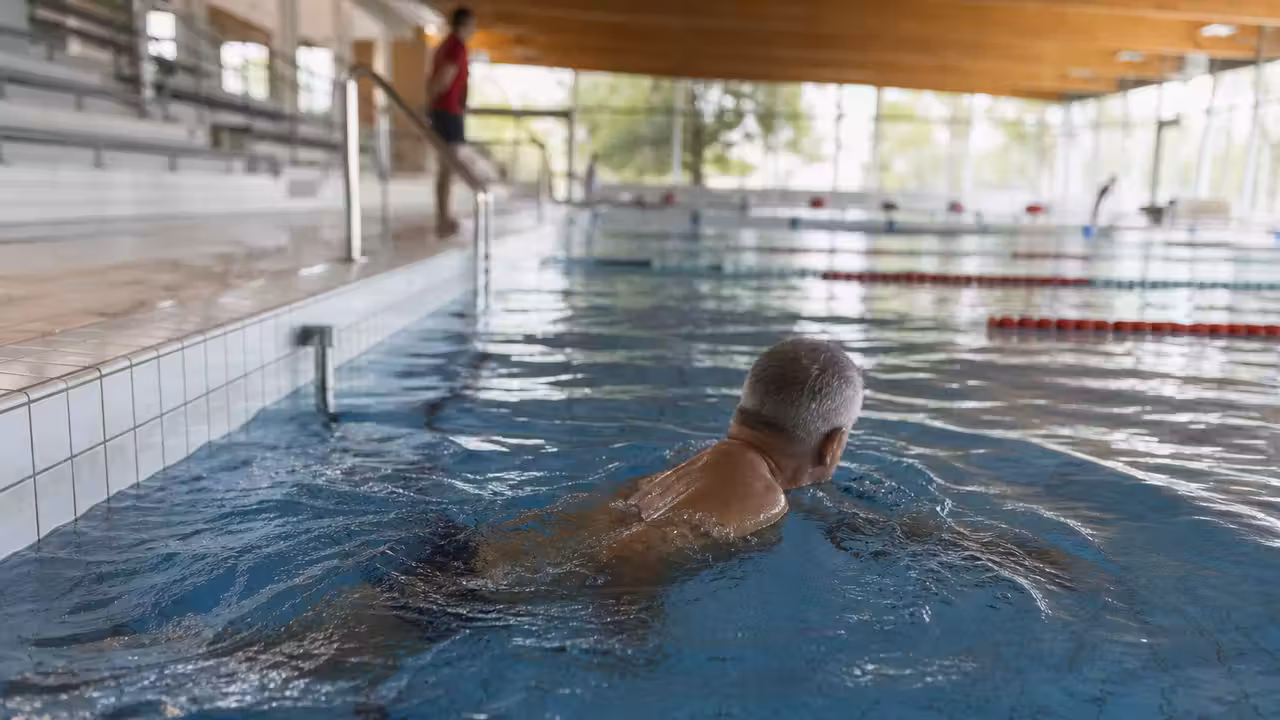 Nageuse senior avançant lentement dans un couloir de piscine intérieure, sous la surveillance d’un maître-nageur