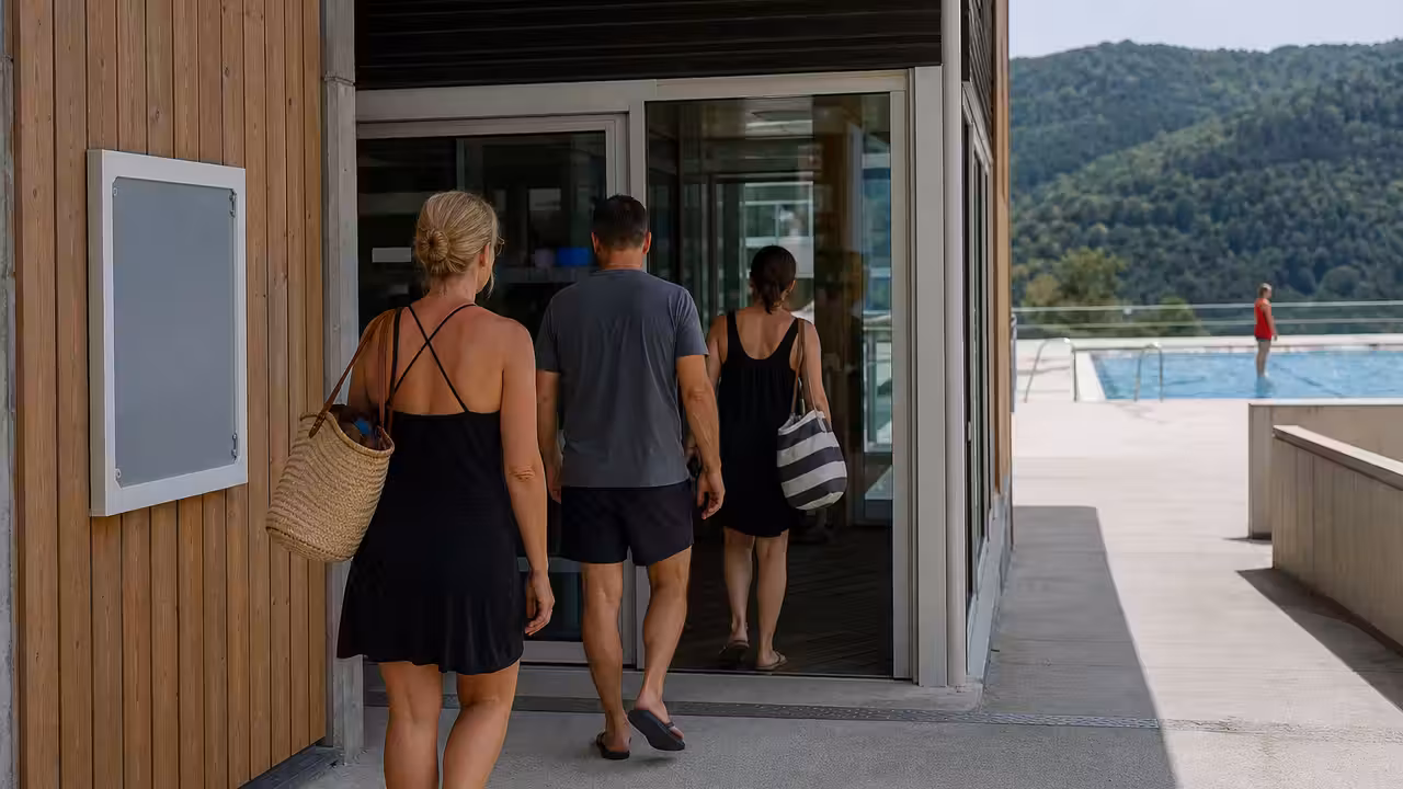 Entrée d’un centre aquatique avec panneau de consignes et bassin extérieur surveillé en arrière-plan