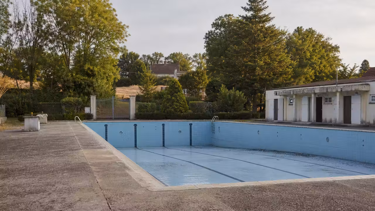 Bassin extérieur municipal vide, gradins et végétation estivale illustrant la fermeture d’une piscine locale