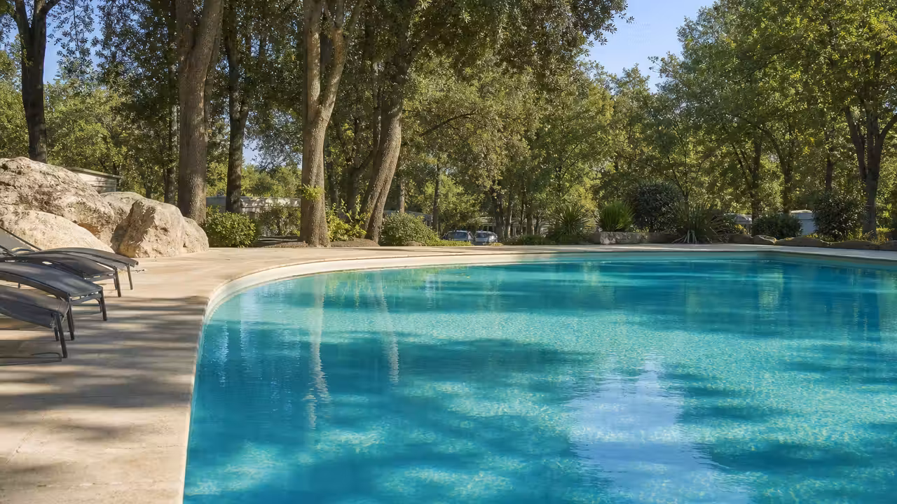 Bassin extérieur d’un camping entouré d’arbres, avec plage minérale et allée piétonne