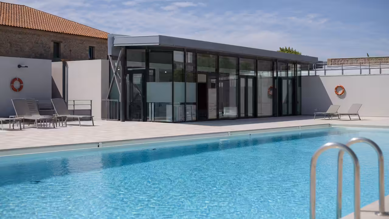 Bassin d’une piscine municipale rénovée avec plages claires, vestiaires vitrés et espace de détente extérieur