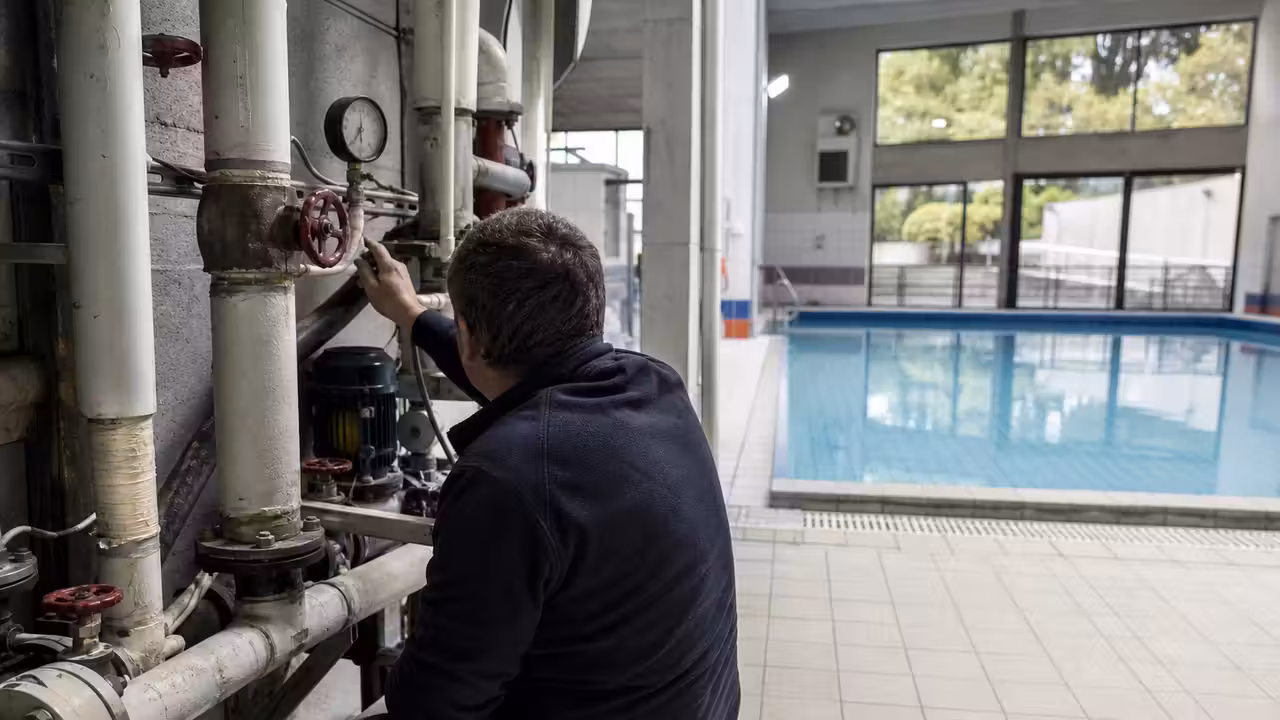 Couloir technique d’une piscine municipale avec canalisations de chauffage et bassin vide en arrière-plan