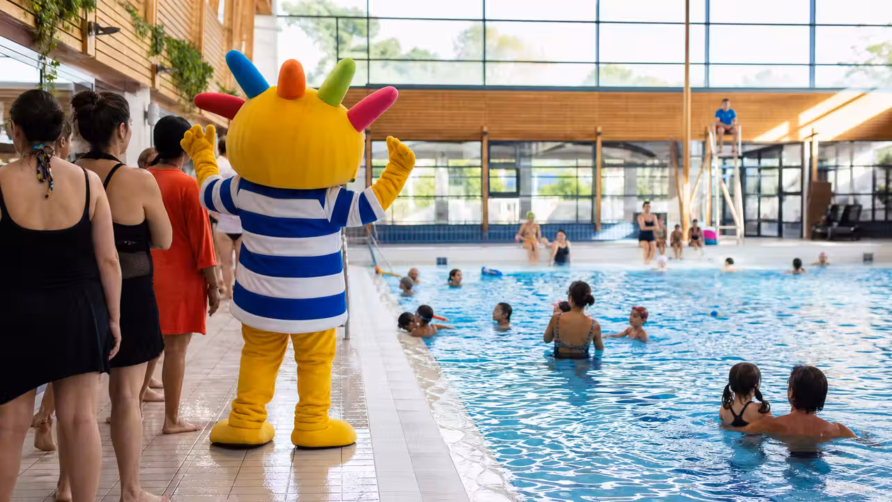 Animation familiale autour d’une mascotte au bord d’un bassin intérieur de centre aquatique municipal