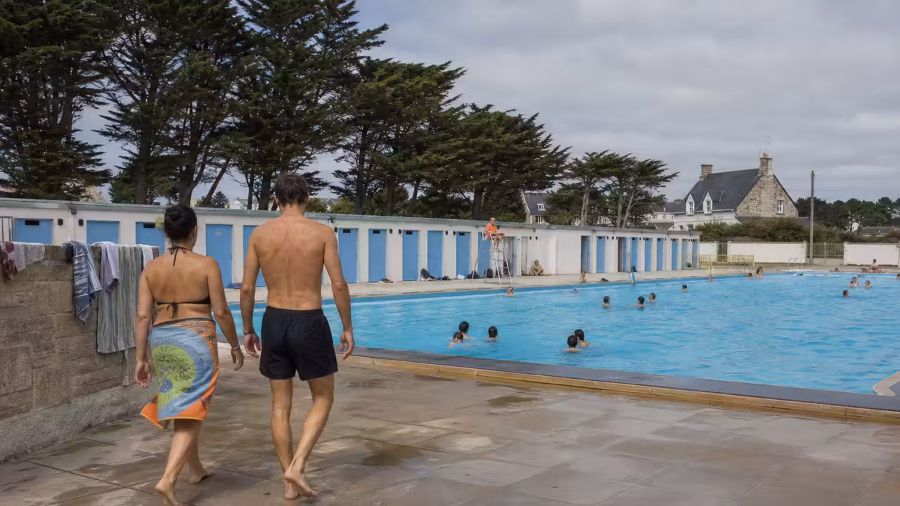Bassin extérieur d’une piscine publique bretonne, baigneurs vus de dos et maître-nageur au loin