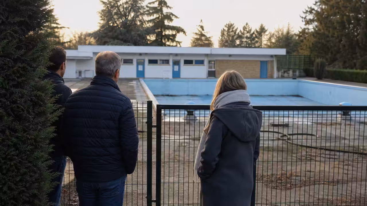 Bassin extérieur municipal vide, grilles fermées et façade technique d’une piscine vieillissante en arrière-plan