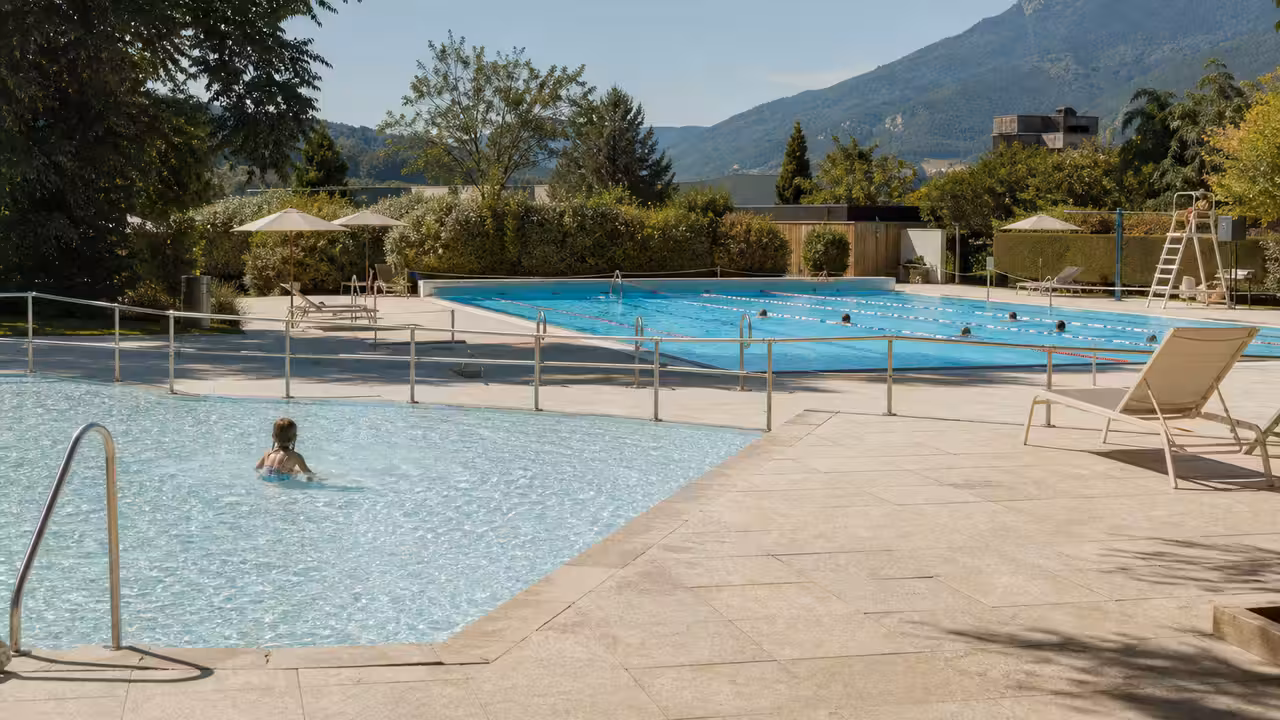 Bassin extérieur municipal bordé de plages minérales et de transats, sous une lumière estivale douce