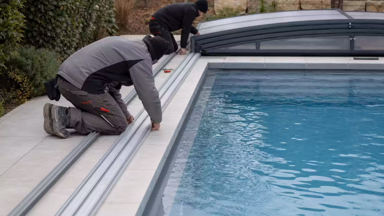 Deux techniciens alignent les rails d’un abri télescopique sur la plage d’une piscine privée.
