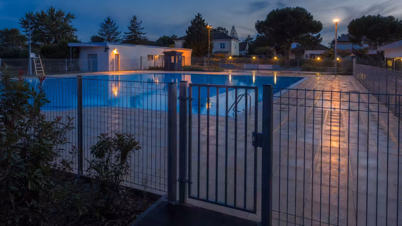 Clôture fermée d’une piscine municipale au crépuscule, avec bassin éclairé mais sans baigneurs