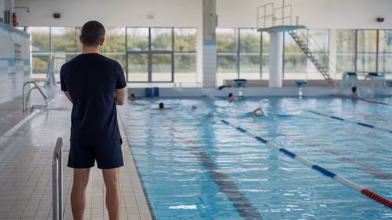 Bassin couvert d’une piscine municipale avec lignes d’eau, baigneurs au loin et maître-nageur au bord