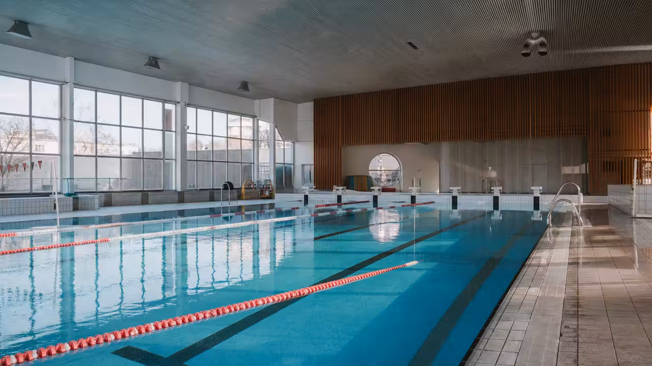 Bassin intérieur d'une piscine municipale vide avec lignes d'eau, gradins discrets et grandes baies vitrées