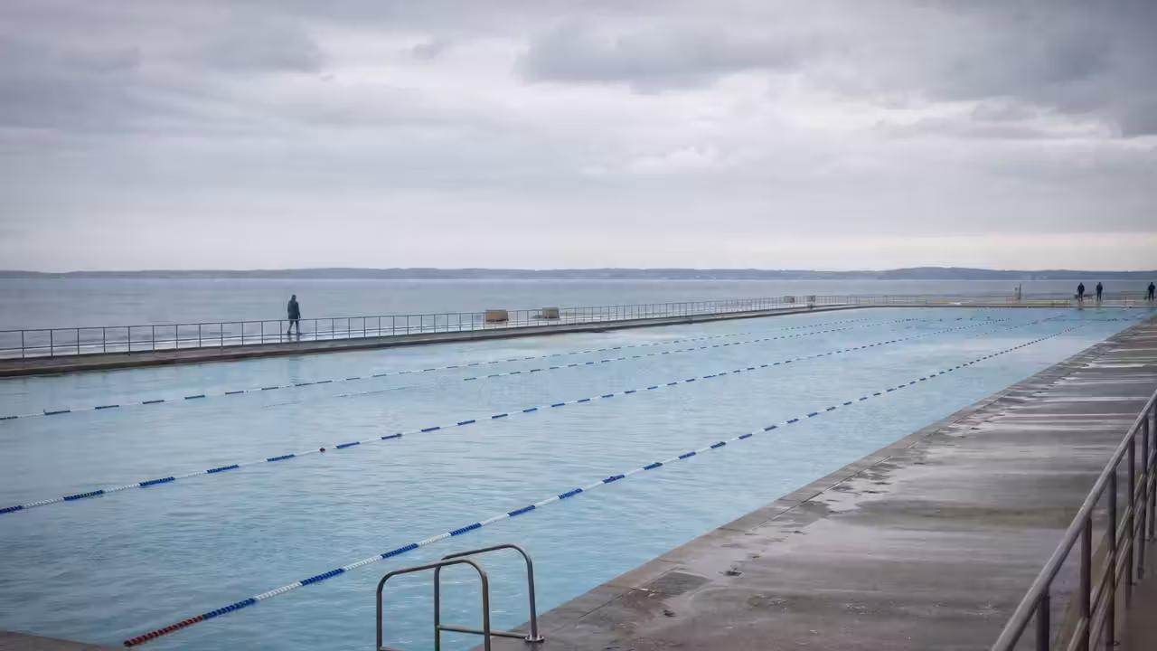 Bassin extérieur de 50 mètres en bord de mer, vide, avec lignes d’eau et gradins sous une lumière hivernale