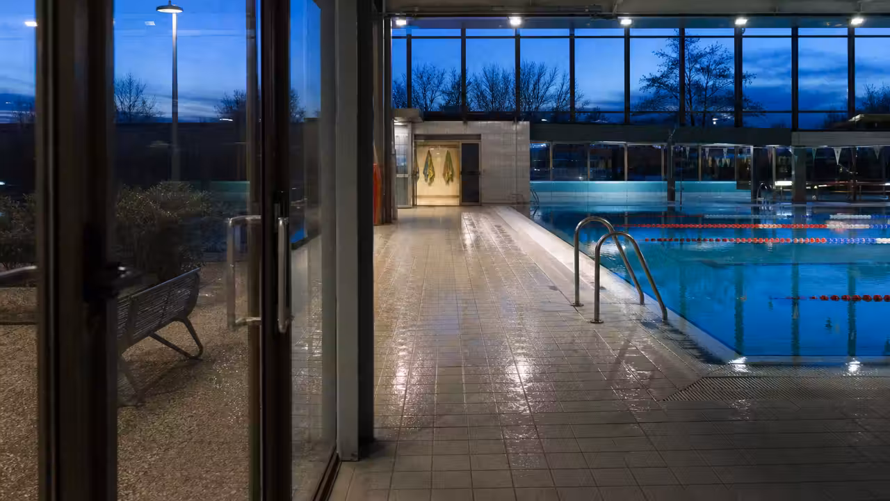 Entrée d’une piscine municipale au crépuscule, avec bassin intérieur éclairé et couloirs de nage vides