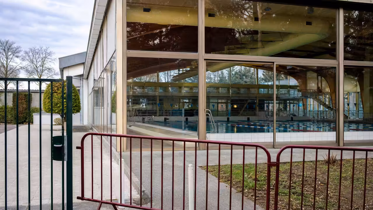 Portail fermé devant une piscine municipale vide, avec bassin couvert et équipements techniques en arrière-plan