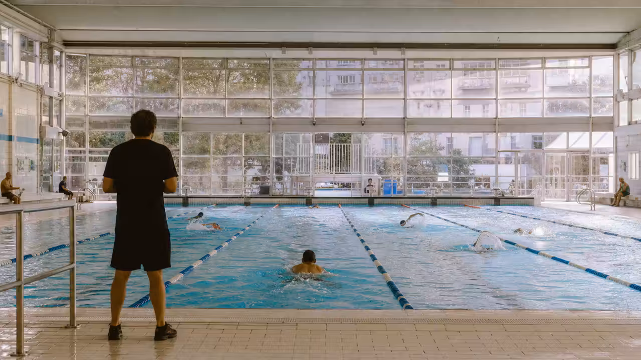 Baigneurs dans un bassin municipal parisien lumineux, avec lignes d’eau et maître-nageur au bord