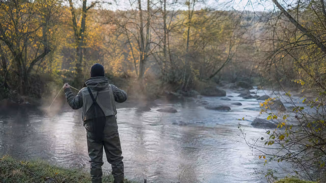 Pêcheur équipé au bord d’une rivière creusoise bordée de rochers et de végétation, au lever du jour