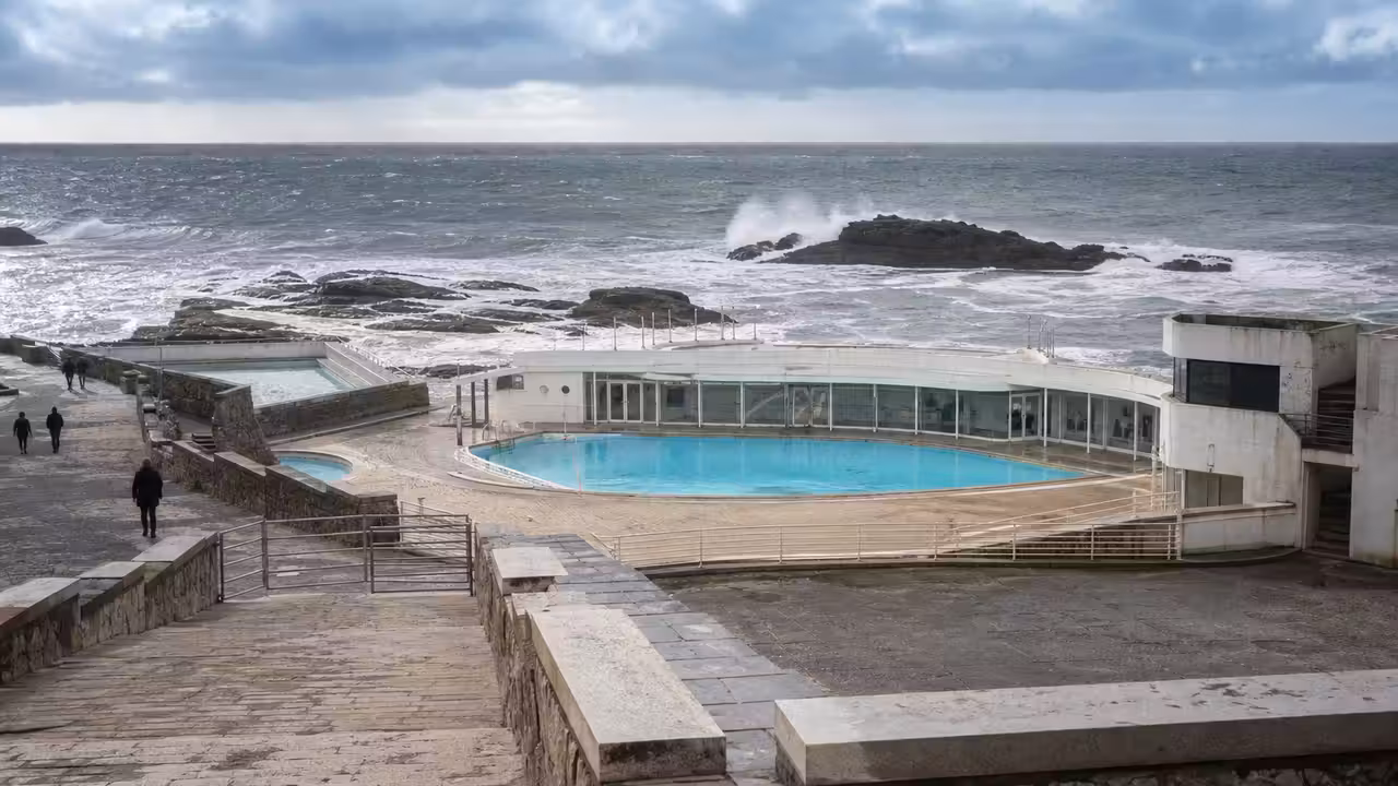 Bâtiment de piscine municipale en front de mer, avec terrasse minérale et vagues à l’arrière-plan