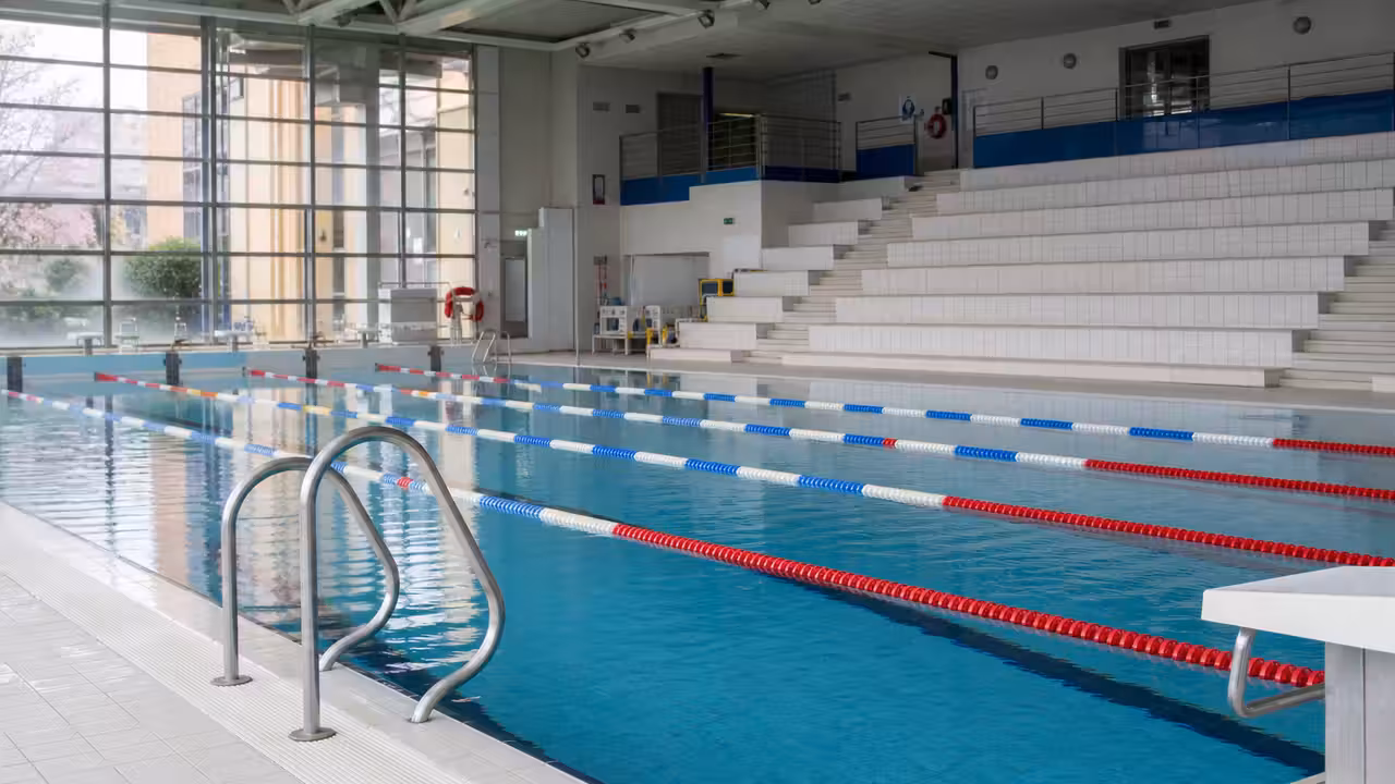 Bassin intérieur d'une piscine municipale avec lignes d'eau, gradins et grandes baies vitrées sans baigneurs visibles