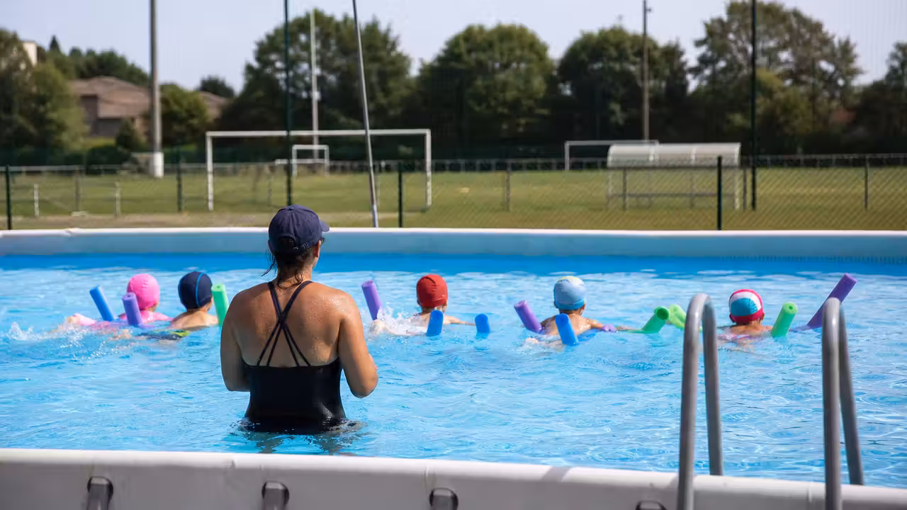 Un maître-nageur accompagne de jeunes enfants vus de dos dans un petit bassin d’apprentissage sécurisé.