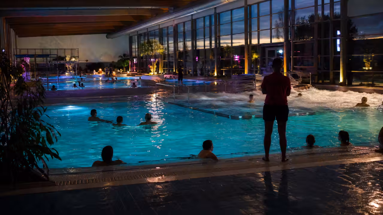 Bassin intérieur éclairé pour une soirée aquatique, avec jeux de lumière et personnel de surveillance en retrait