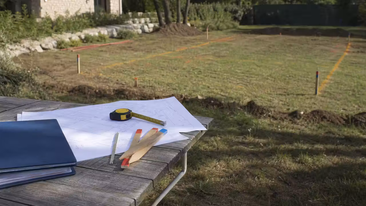 Plan de masse, mètre ruban et dossier d’urbanisme posés près d’un terrain préparé pour une piscine