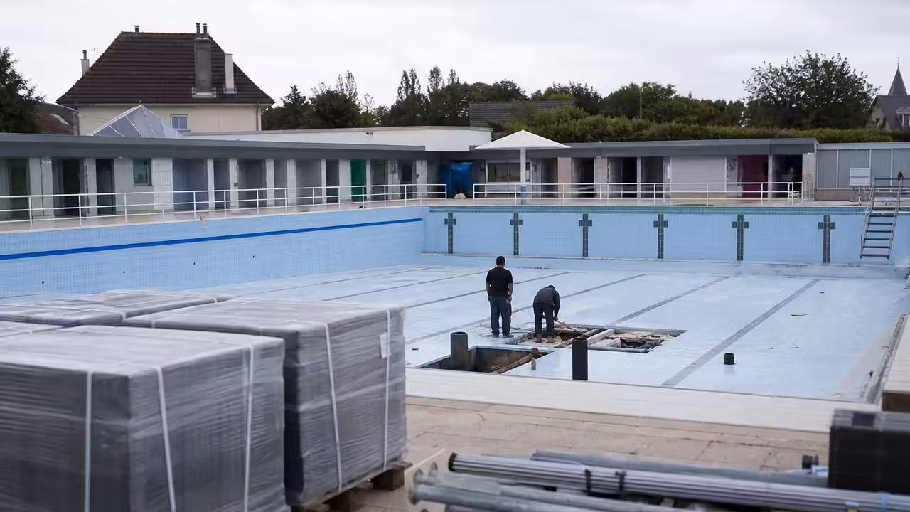 Bassin municipal vide en rénovation, avec plages carrelées et équipements techniques en arrière-plan