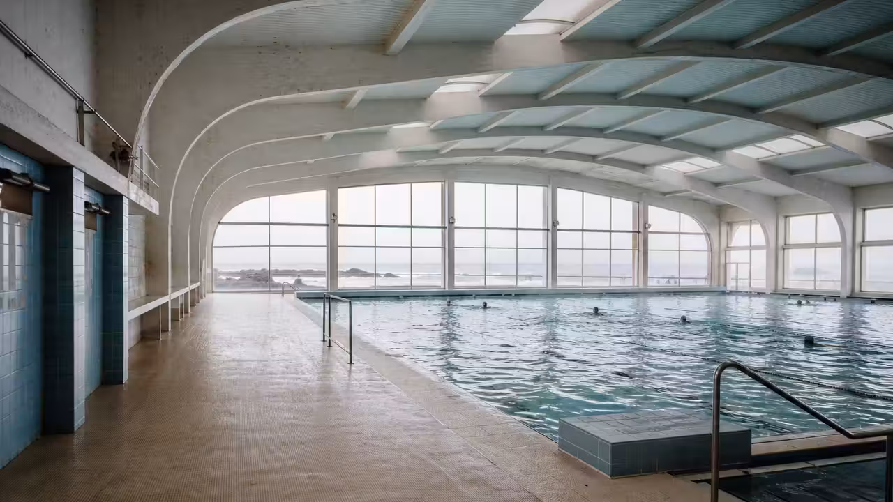 Bassin couvert municipal à l’architecture des années 1960, avec nageurs vus de loin et carrelage bleu