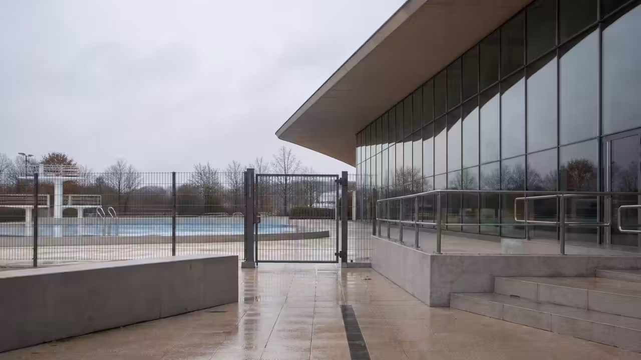 Grilles fermées devant un bassin extérieur municipal vide, symbole d’un équipement aquatique arrêté