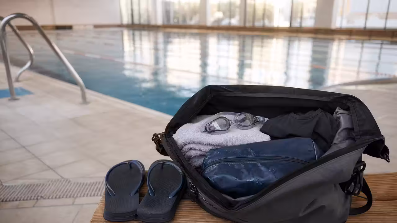 Sac de piscine ouvert sur un banc avec serviette, lunettes, maillot et pochette pour linge humide