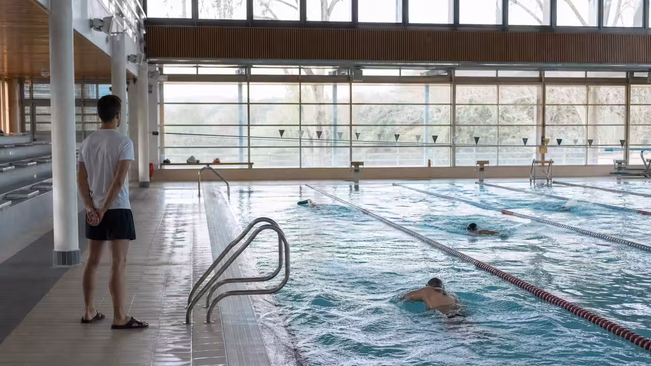 Couloirs d’une piscine municipale éclairée le matin, avec quelques nageurs adultes et un maître-nageur au bord
