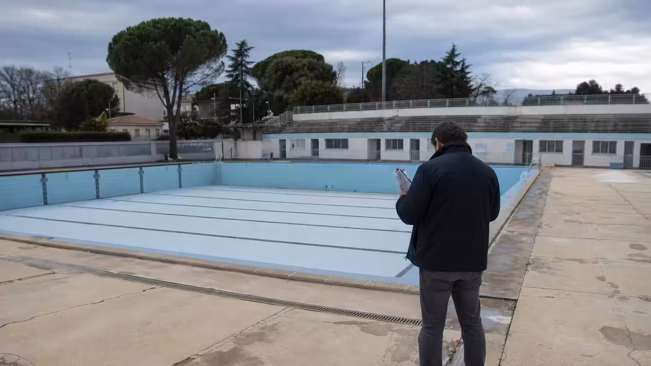 Plages en béton légèrement fissurées autour d’un bassin extérieur de piscine publique vide, avec caniveaux visibles