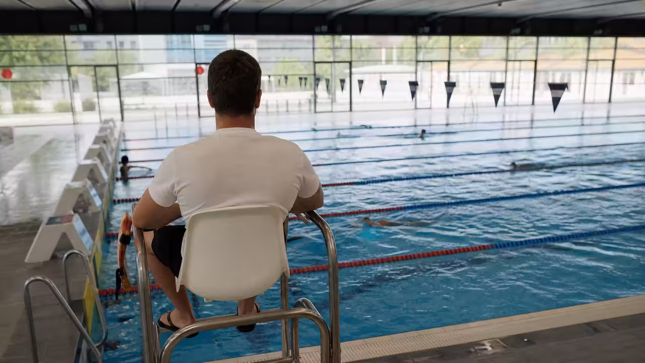Maître-nageur surveillant un bassin couvert depuis son poste, avec plusieurs lignes d’eau occupées