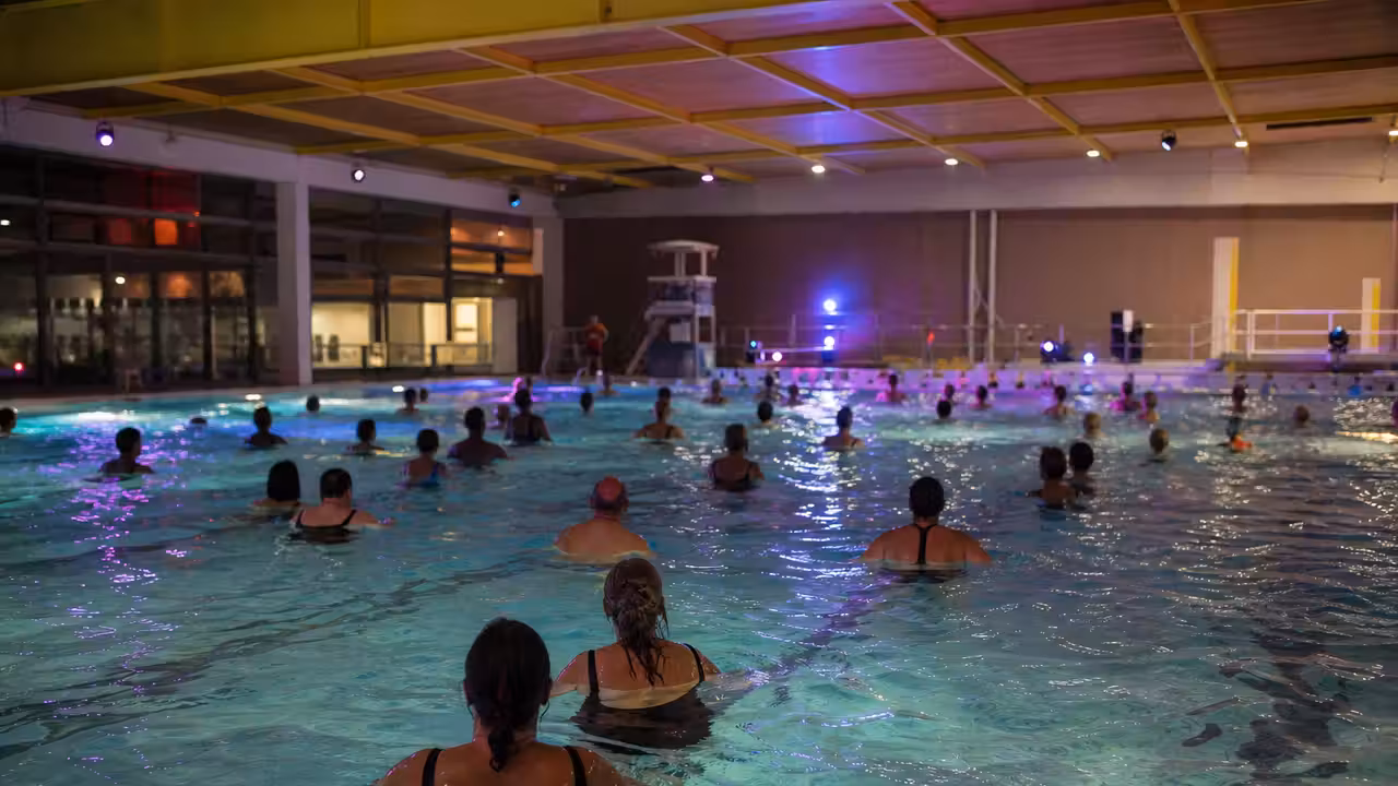 Bassin municipal illuminé pour une soirée aquatique, avec participants de dos dans une ambiance festive