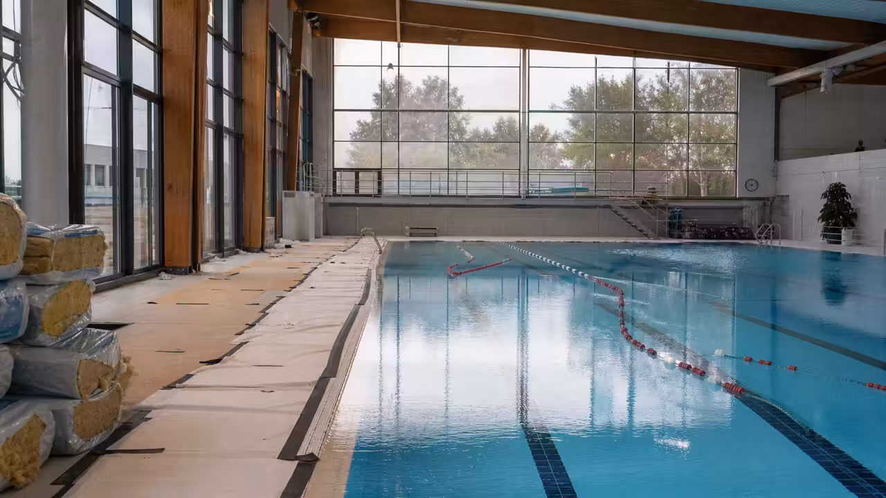 Hall de bassin couvert vide pendant des travaux d’isolation, avec lignes d’eau et baies vitrées en arrière-plan
