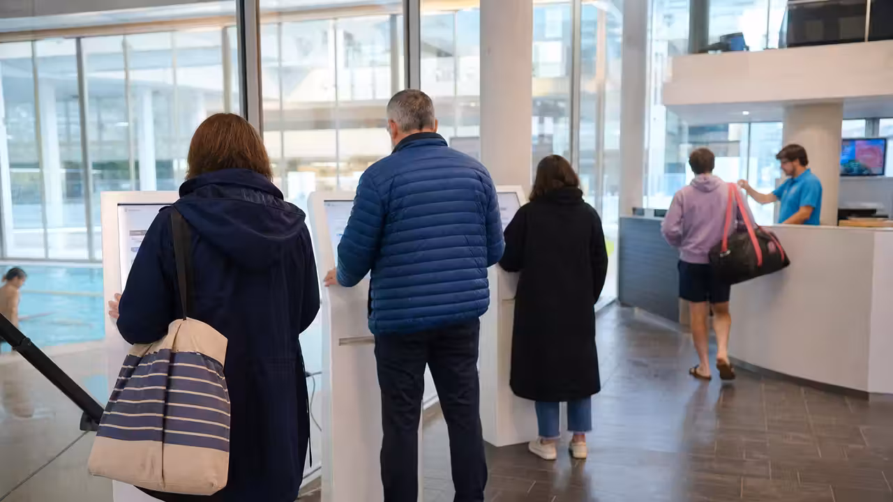 Usagers devant des bornes de billetterie à l’entrée d’une piscine municipale lumineuse et contemporaine