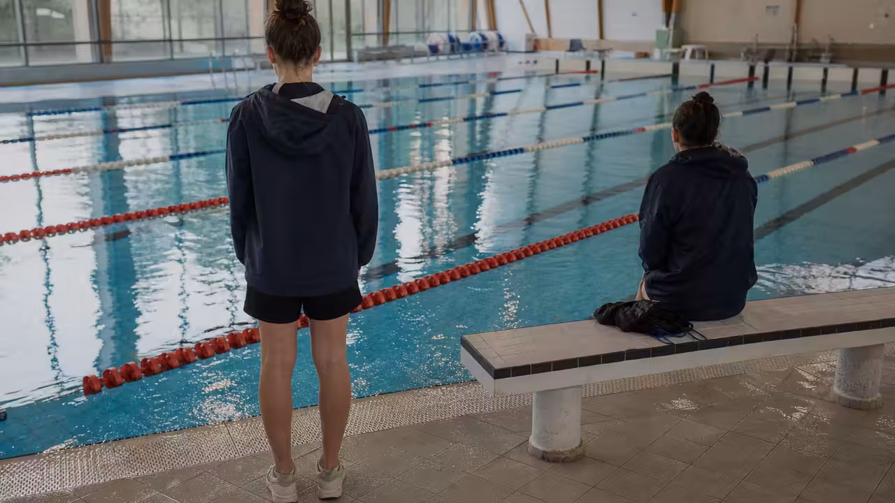 Deux jeunes nageurs vus de dos au bord d'un bassin couvert, dans une atmosphère calme et introspective.