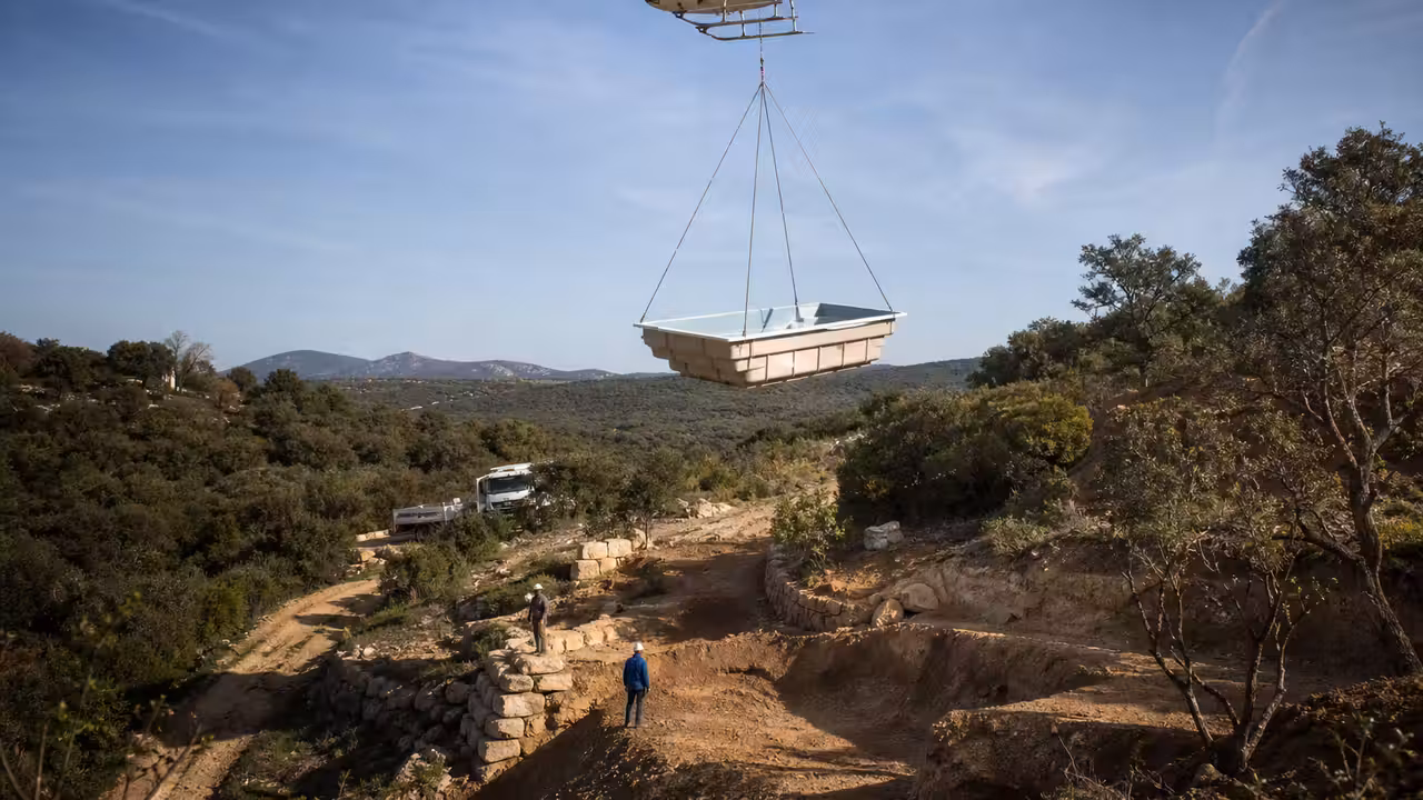 Piscine coque suspendue sous un hélicoptère au-dessus d’un terrain rural difficilement accessible