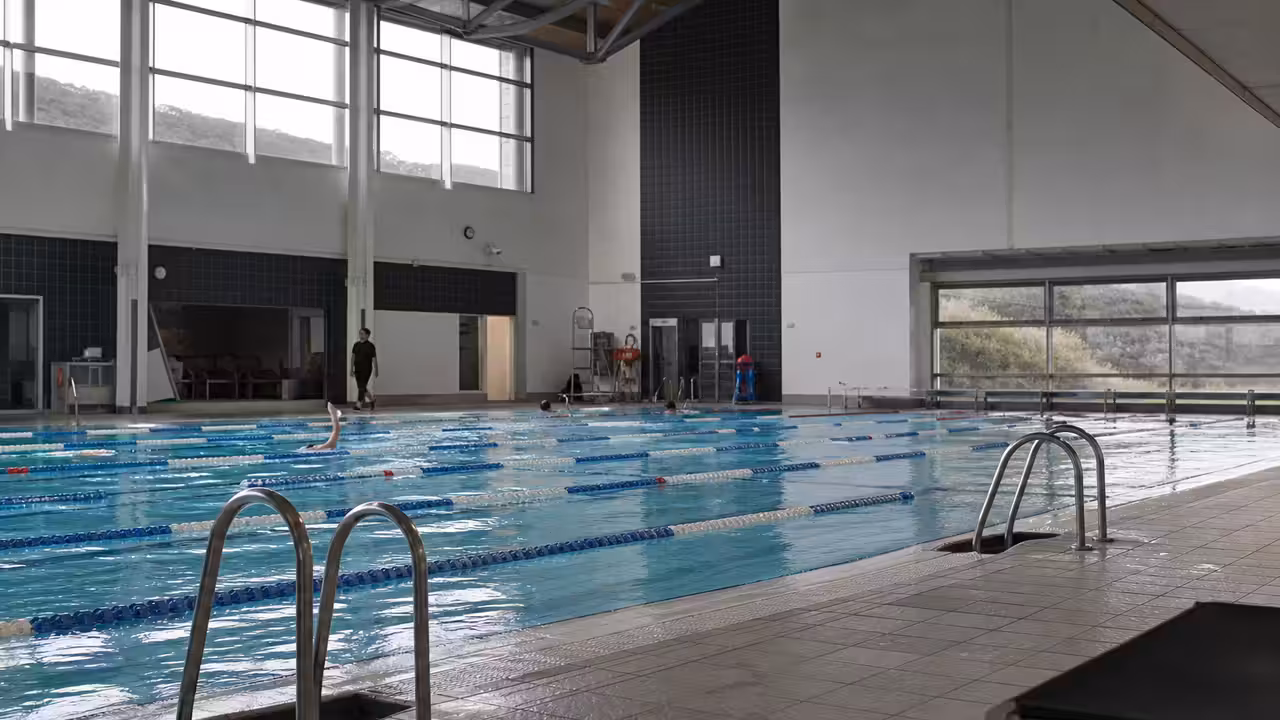 Bassin couvert d’un centre aquatique avec lignes de nage, baies vitrées et nageurs de dos au loin