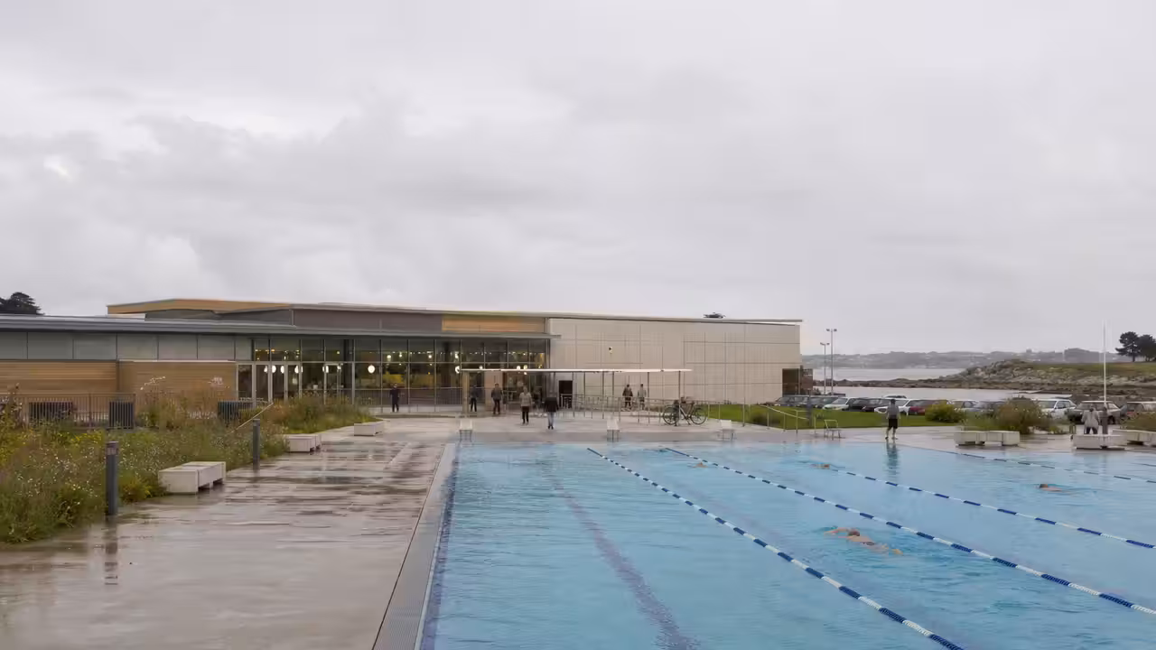 Vue extérieure d’un centre aquatique municipal moderne avec bassin extérieur de 25 mètres près d’équipements sportifs.