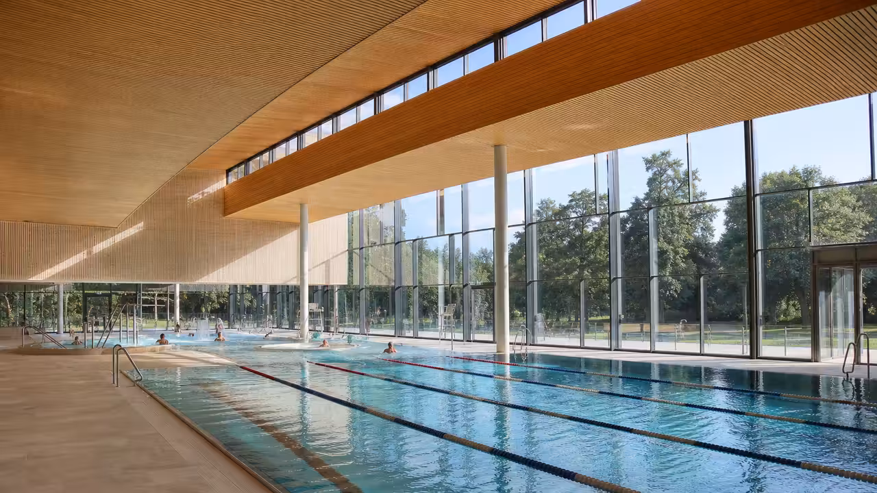 Vue intérieure lumineuse d’un centre aquatique municipal avec grand bassin, baies vitrées et nageurs éloignés