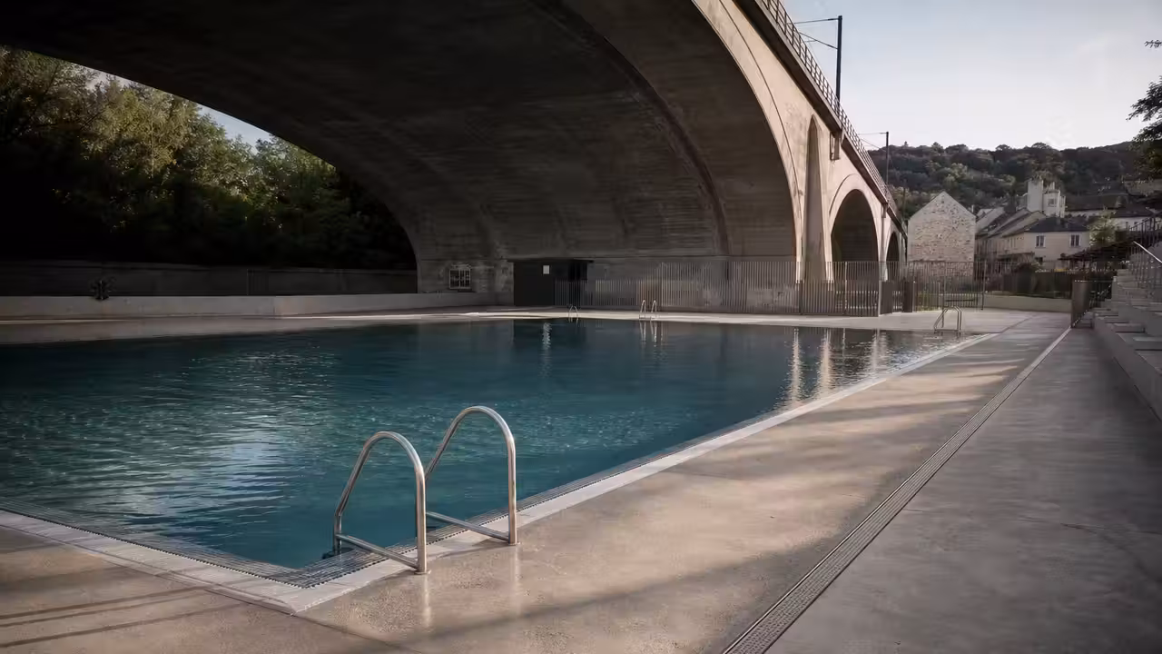 Bassin de baignade urbain installé sous un pont ferroviaire, avec plages minérales et lumière naturelle latérale