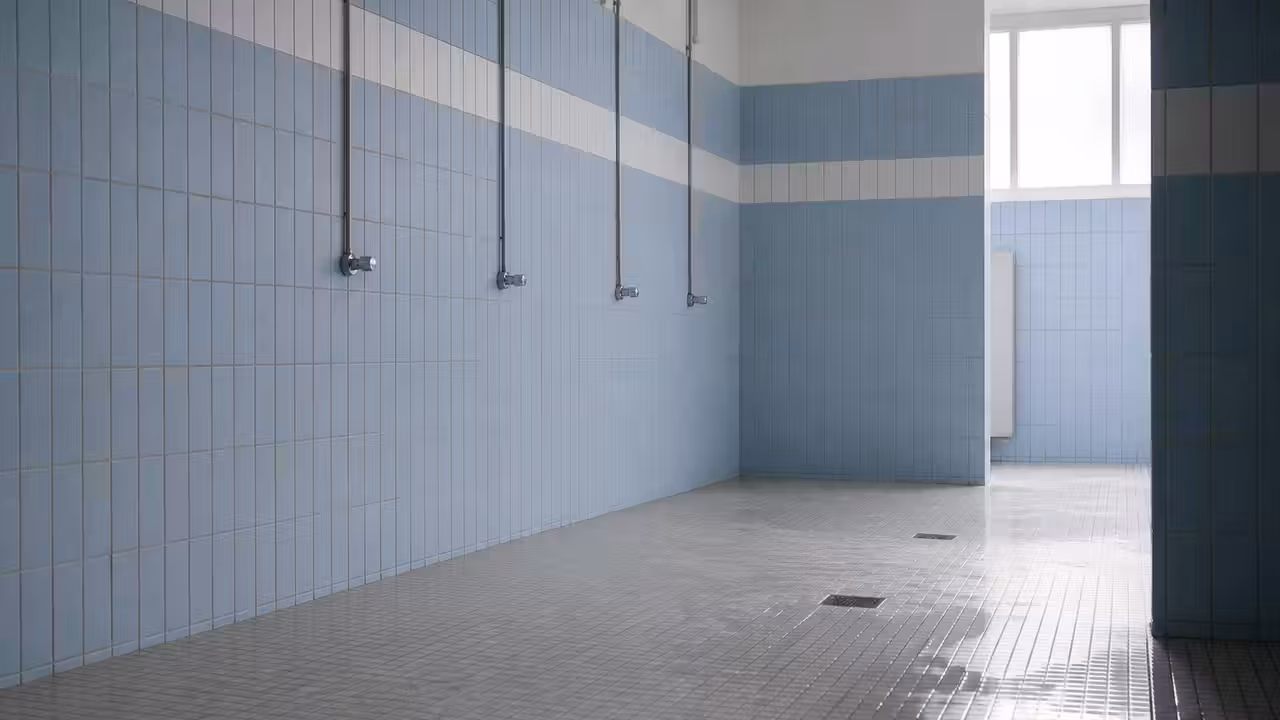 Vestiaire de piscine municipale avec rangée de douches, sol carrelé et évacuations au premier plan