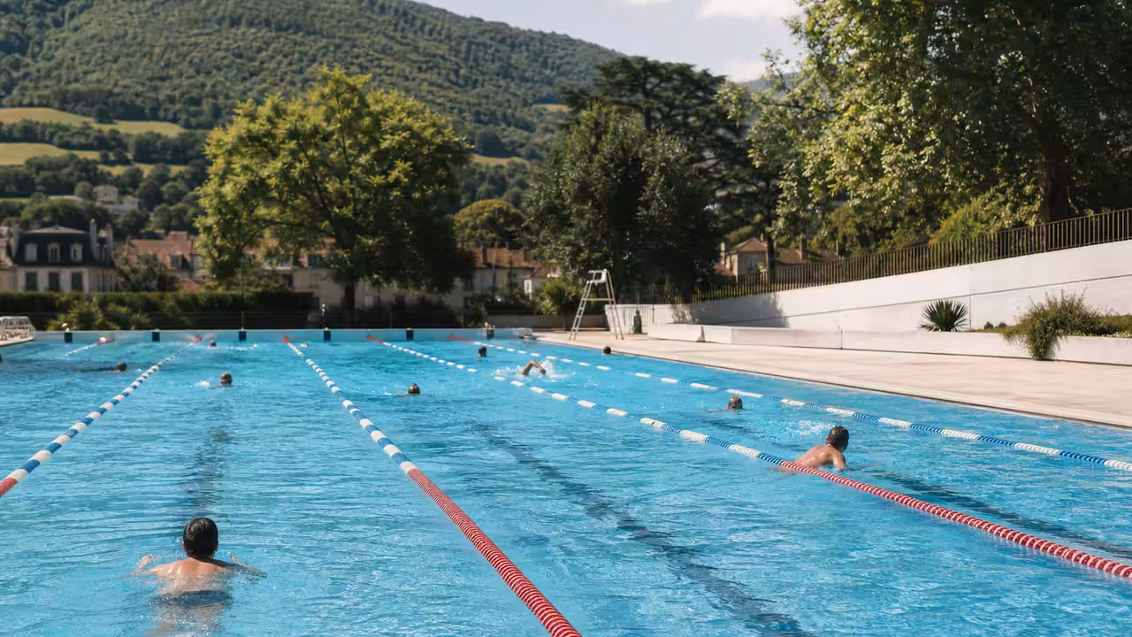 Bassin extérieur municipal avec lignes d’eau, plages minérales et baigneurs vus de dos par une journée d’été